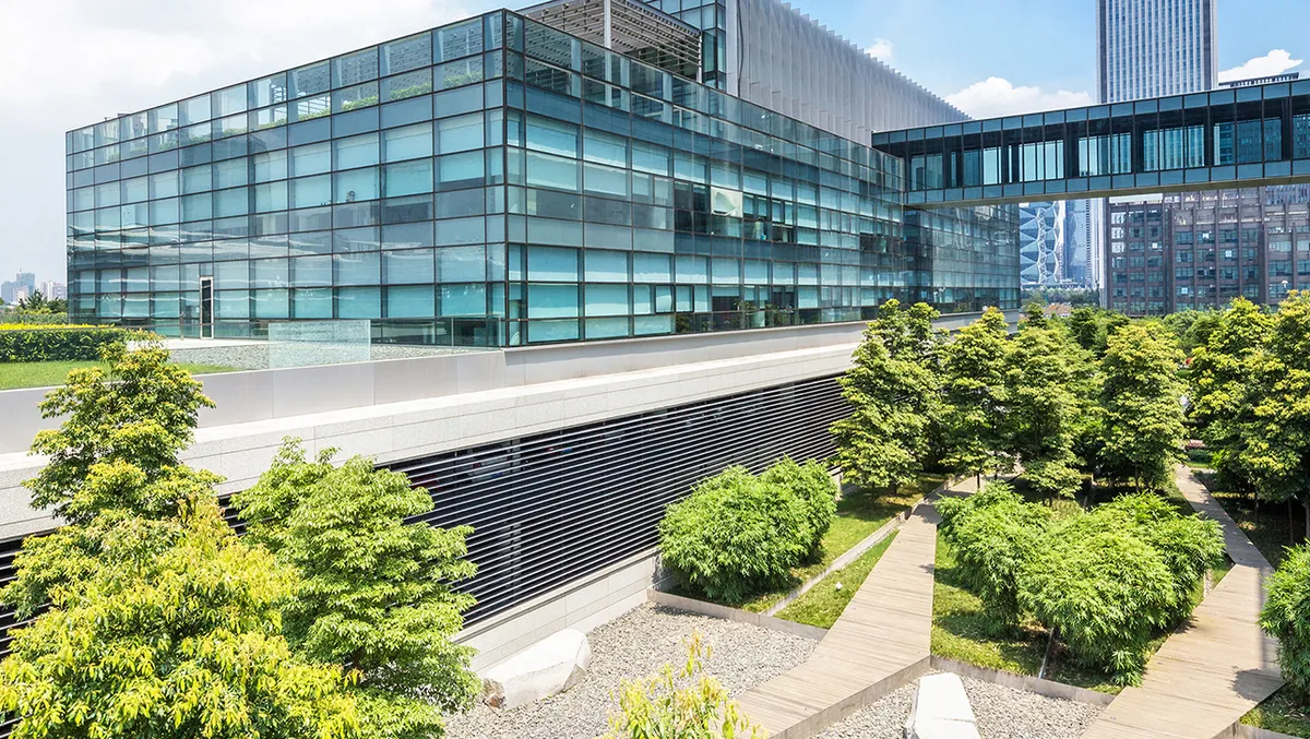 Modernes Bürogebäude mit Glasfassade und begrüntem Außenbereich. Vor dem Gebäude befinden sich gepflegte Bäume, Sträucher und ein Holzsteg, der sich durch die Anlage schlängelt. Eine gläserne Brücke verbindet die Gebäudeteile, im Hintergrund sind weitere Hochhäuser der Stadt zu sehen. Modernes Bürogebäude mit Glasfassade und begrüntem Außenbereich. Vor dem Gebäude befinden sich gepflegte Bäume, Sträucher und ein Holzsteg, der sich durch die Anlage schlängelt. Eine gläserne Brücke verbindet die Gebäudeteile, im Hintergrund sind weitere Hochhäuser der Stadt zu sehen.