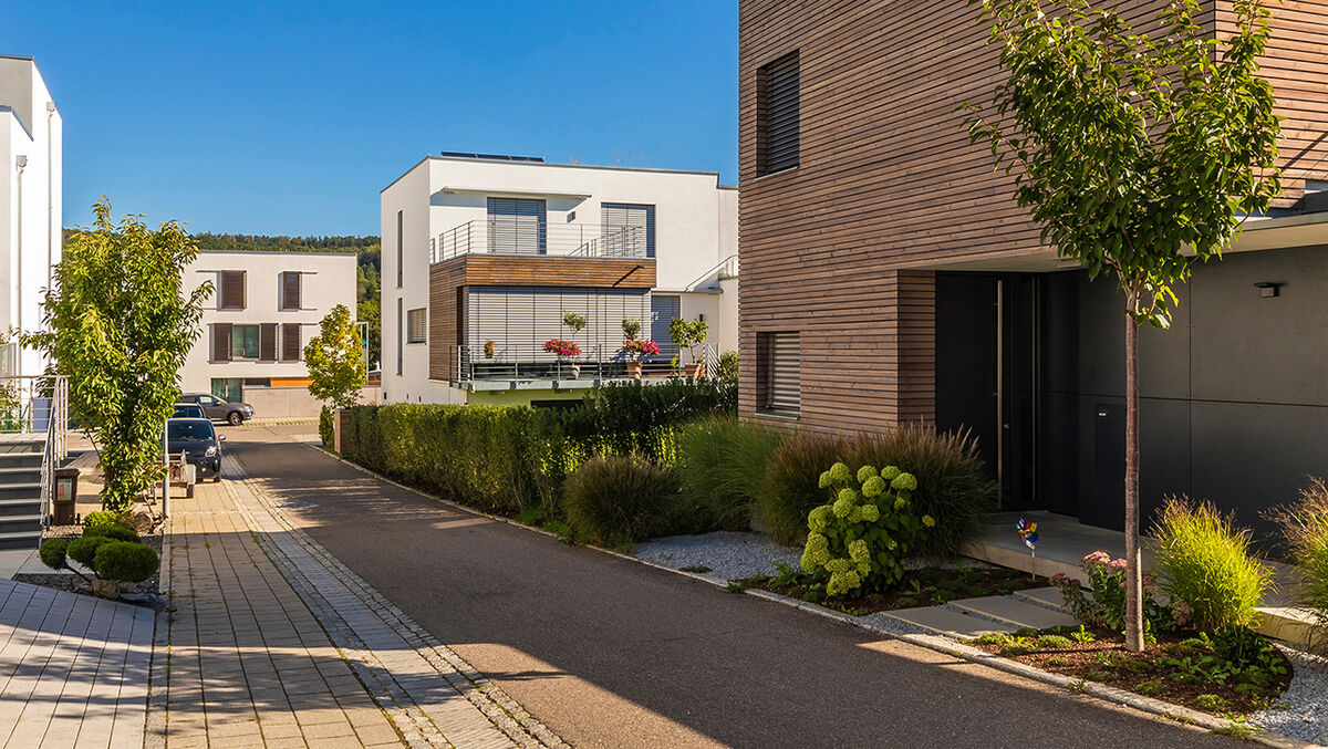 Ruhige Wohnstraße mit modernen Einfamilienhäusern in kubischer Architektur. Die Gebäude haben helle Fassaden, teils mit Holzverkleidung und großen Fenstern. Begrünte Vorgärten, Bäume und Pflanzbeete verleihen dem Viertel eine gepflegte und naturnahe Atmosphäre bei sonnigem Wetter. Ruhige Wohnstraße mit modernen Einfamilienhäusern in kubischer Architektur. Die Gebäude haben helle Fassaden, teils mit Holzverkleidung und großen Fenstern. Begrünte Vorgärten, Bäume und Pflanzbeete verleihen dem Viertel eine gepflegte und naturnahe Atmosphäre bei sonnigem Wetter.