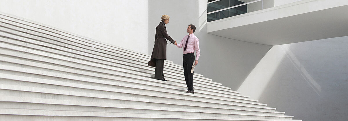 A woman and a man in business attire stand on a large, bright staircase of an office building, smiling and shaking hands