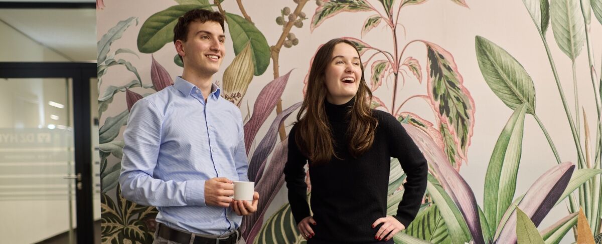 Zwei junge Personen stehen nebeneinander und lächeln. Hinter ihnen befindet sich eine Wand mit floralem Muster. Zwei junge Personen stehen nebeneinander und lächeln. Hinter ihnen befindet sich eine Wand mit floralem Muster.
