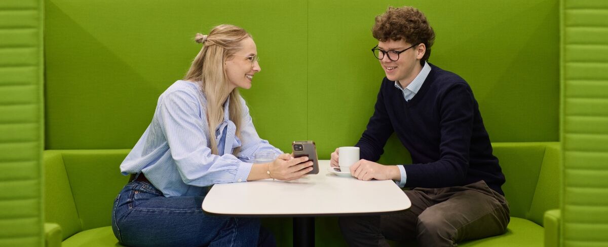 Zwei Personen sitzen sich in einer grünen Sitznische an einem Tisch gegenüber. Sie lächeln und schauen gemeinsam auf ein Smartphone. Zwei Personen sitzen sich in einer grünen Sitznische an einem Tisch gegenüber. Sie lächeln und schauen gemeinsam auf ein Smartphone.