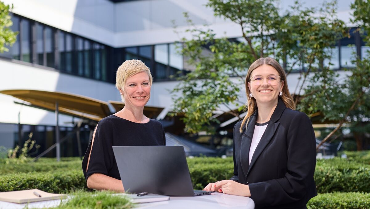 Stellenangebote Zwei Frauen stehen in einem Innenhof an einem Tisch und lächeln in die Kamera. Vor ihnen liegt ein geöffneter Laptop.