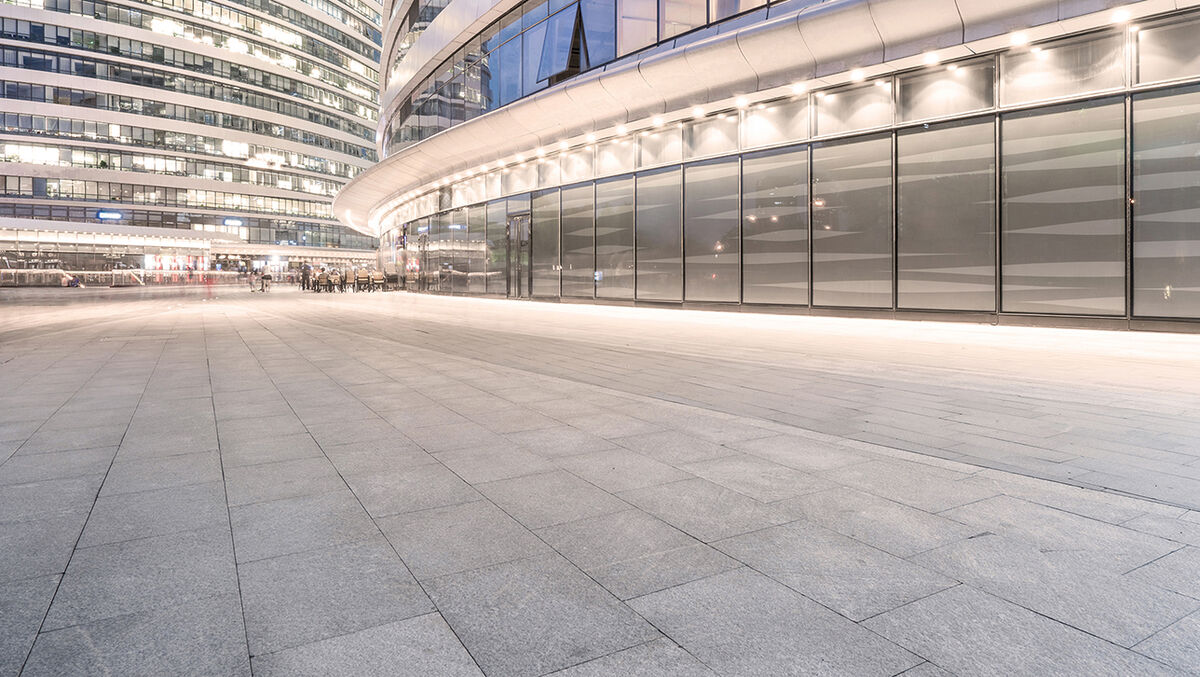Real estate financing for corporate customers Weitwinkelaufnahme eines modernen Büro- oder Geschäftsgebäudes mit geschwungener Glasfassade und weitläufigem, hell gepflastertem Vorplatz. Die Szene ist abends aufgenommen, Innenbeleuchtung und Fassadenlichter sorgen für eine elegante Atmosphäre.